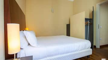 A neatly made bed with a white comforter and pillows, a bedside lamp, and a wooden floor.