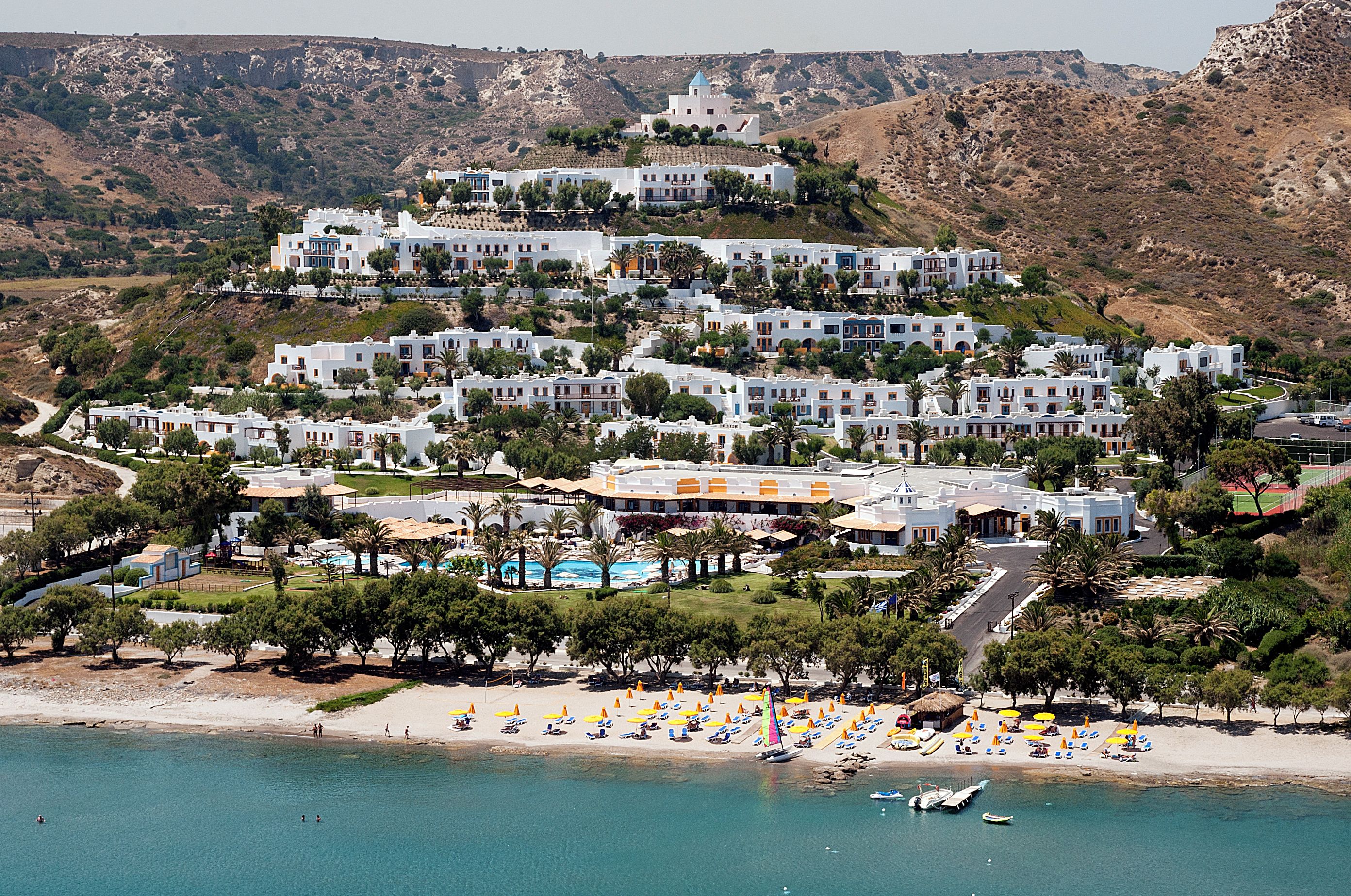 A coastal resort with multiple white buildings, a swimming pool, and a beach with umbrellas.