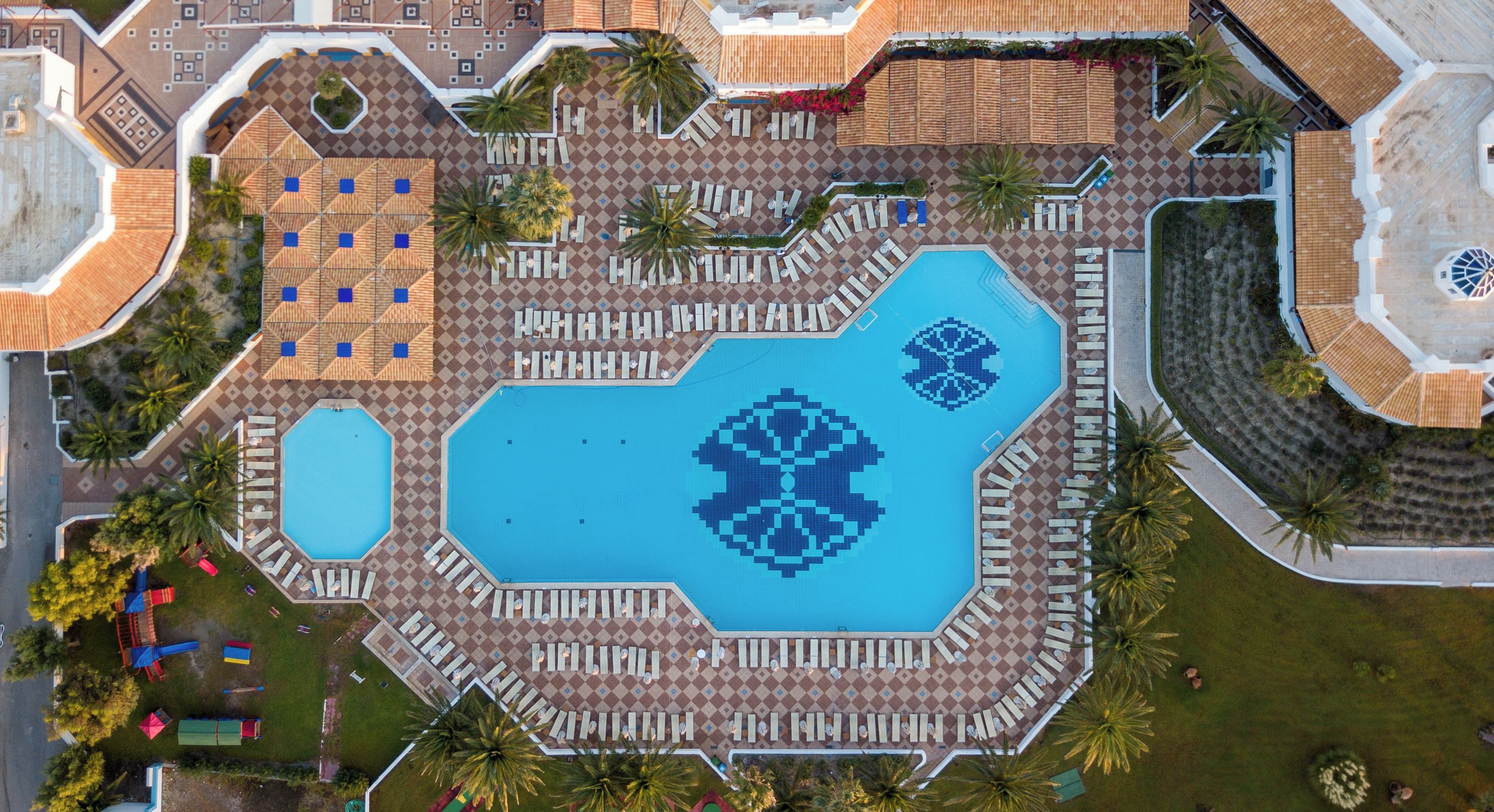 An aerial view of a resort with a central swimming pool, multiple lounge areas, and palm trees.