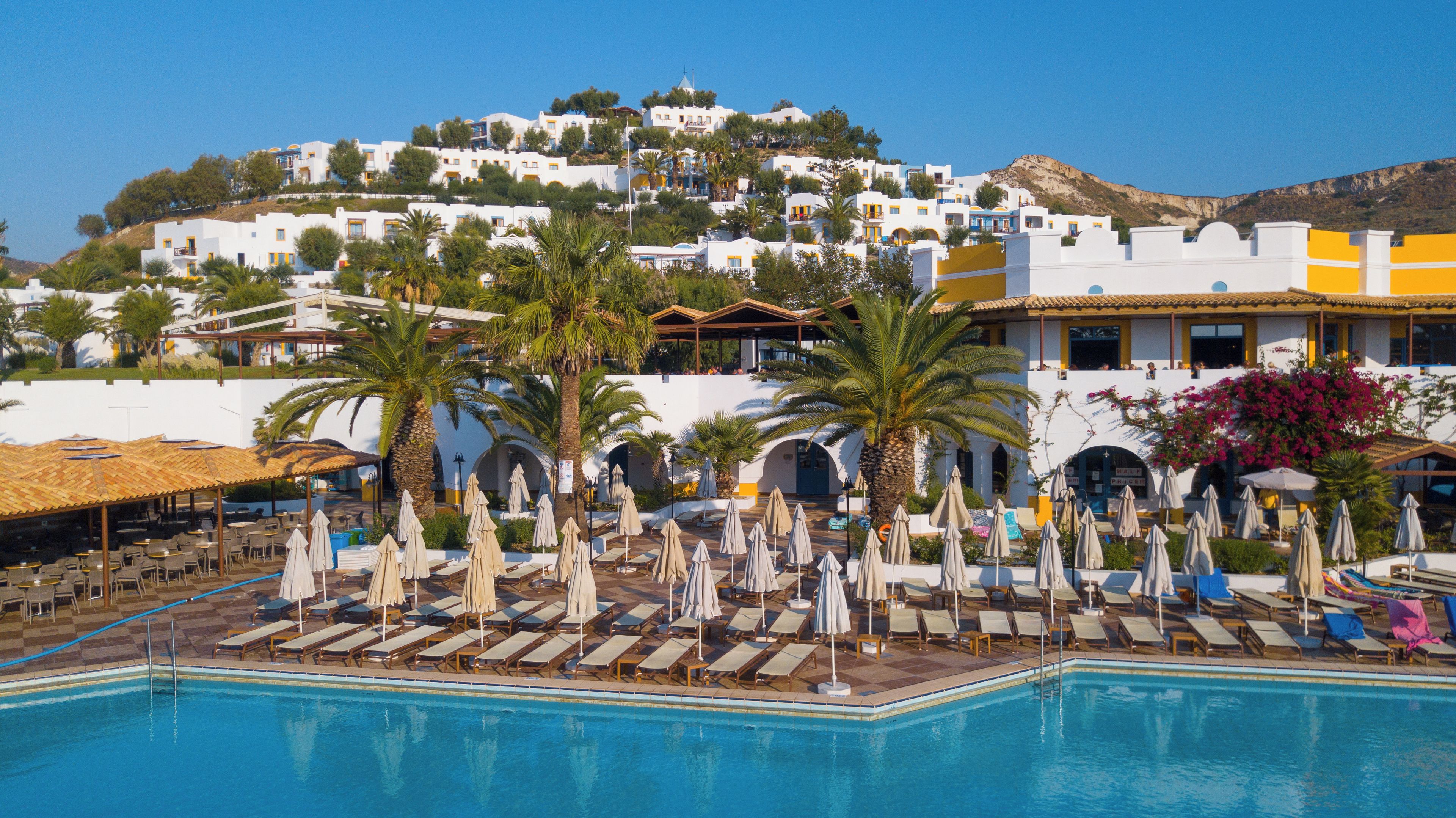 A resort with a pool, sun loungers, umbrellas, and a view of white buildings on a hill.