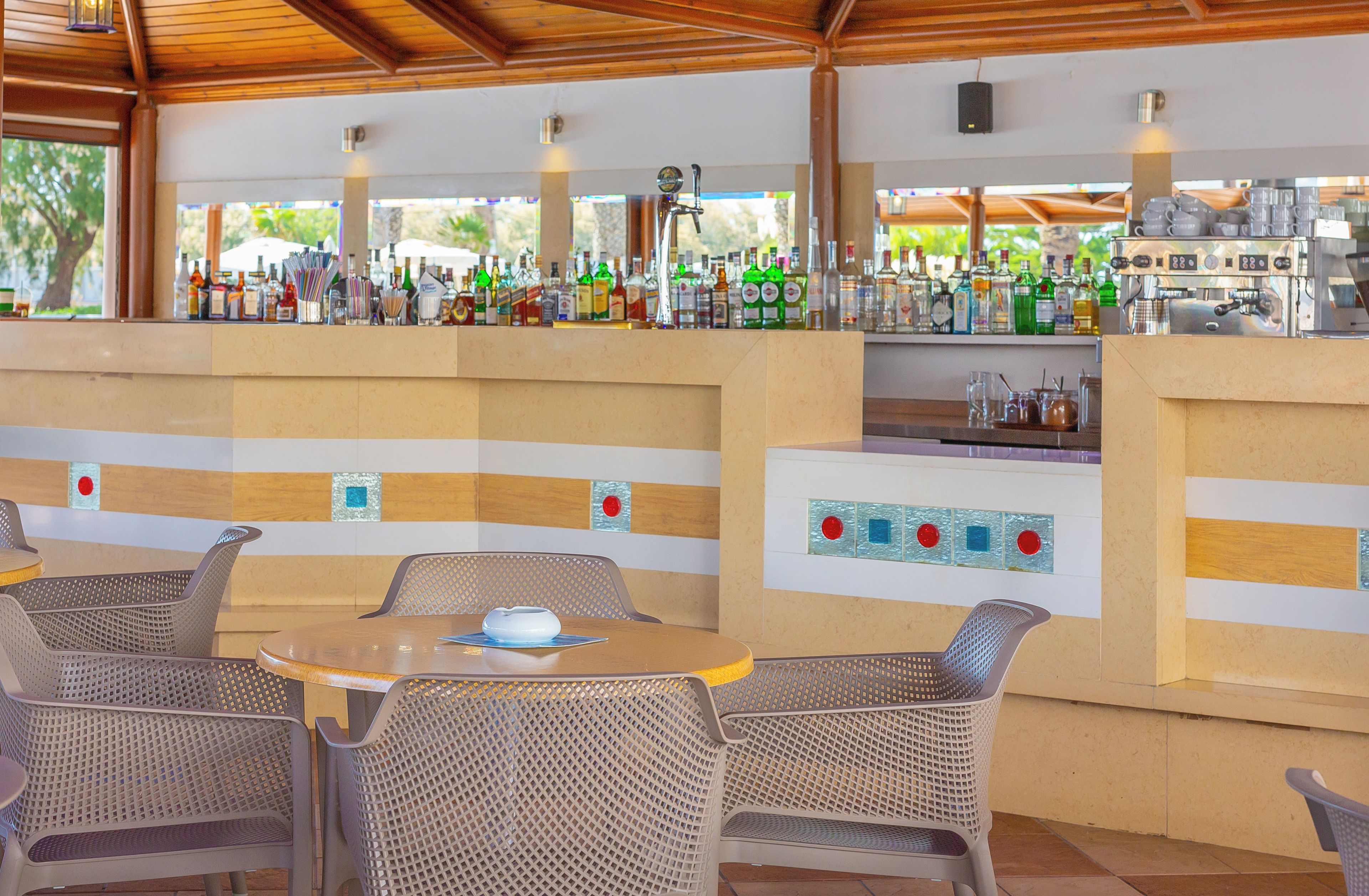 Área de bar externa com cadeiras de vime e uma mesa redonda, balcão de bar com diversos frascos, e vista para o verde através de grandes janelas.
