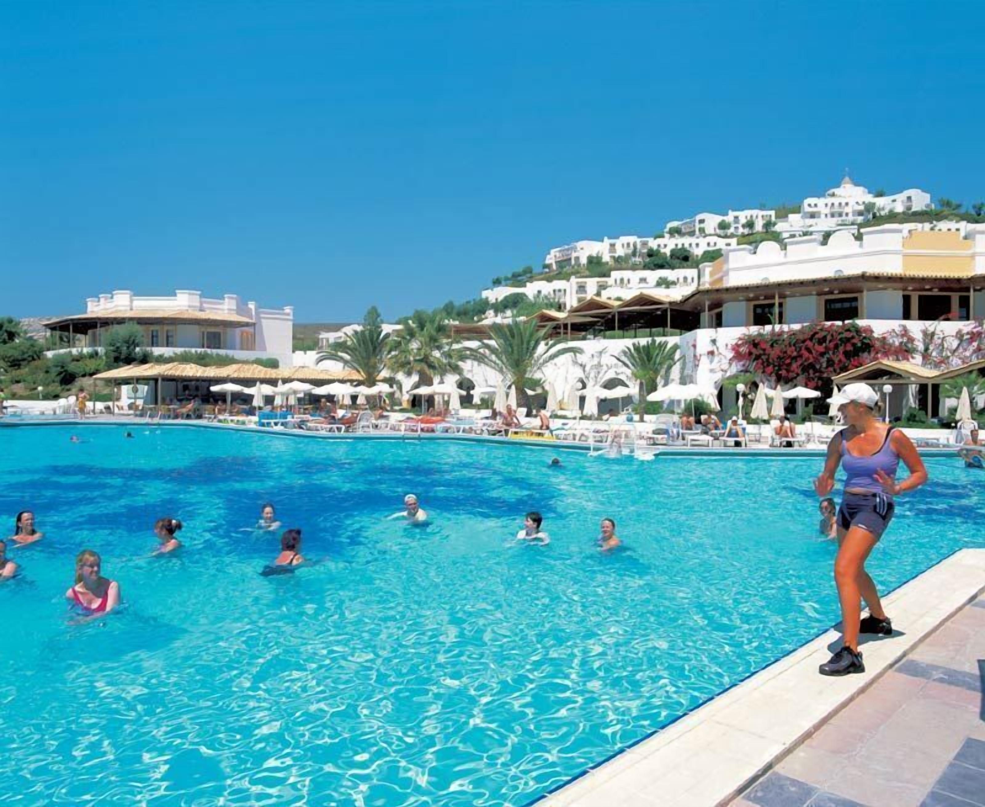 A swimming pool with people in it, surrounded by white buildings and palm trees.