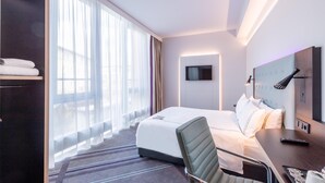 Habitació d'hotel amb un llit, escriptori, cadira, televisió i una finestra gran amb cortines.