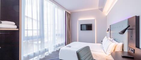 Habitación de hotel con cama, escritorio, silla, televisión y un amplio ventanal con cortinas.