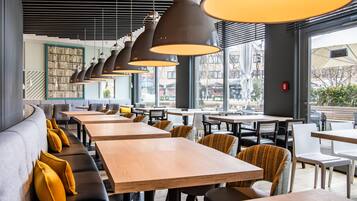 A restaurant with long wooden tables and black chairs, large windows, and hanging pendant lights.