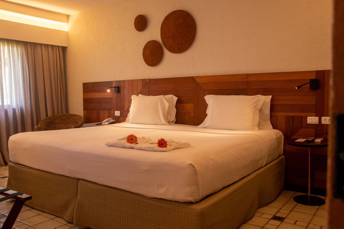 A hotel room with a large bed, wooden headboard, white bedding, a small bedside table, and a wall-mounted lamp.