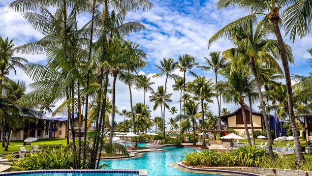A pool surrounded by palm trees and lounge chairs, with a building in the background.