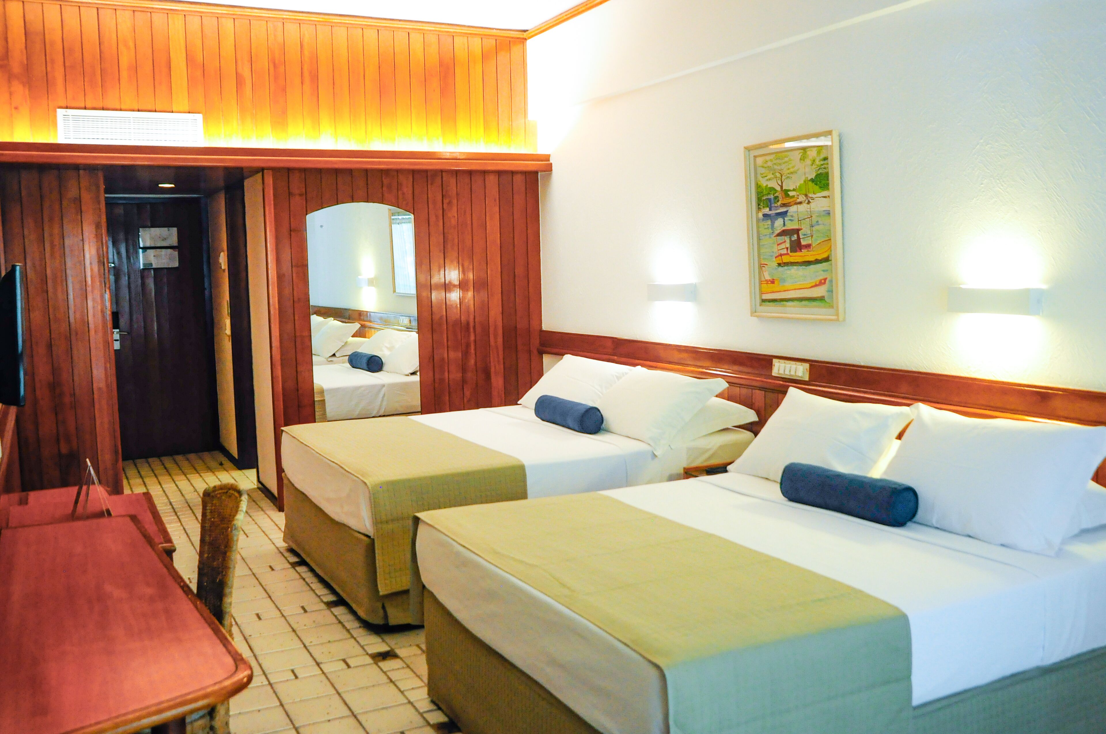Habitación de hotel con dos camas, cabecera de madera, puerta de madera, televisor y un cuadro en la pared.