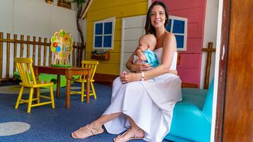Uma mulher sentada em um sofá azul numa sala com cerca de madeira, uma mesa de madeira e uma casinha de brincar colorida ao fundo.