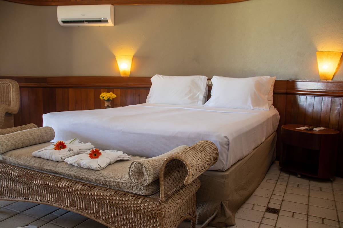 A neatly made bed with white linens, a wicker chair with towels, and a small table with a lamp.