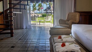 A hotel room with a bed, a wicker chair, and a view of the beach and umbrellas.