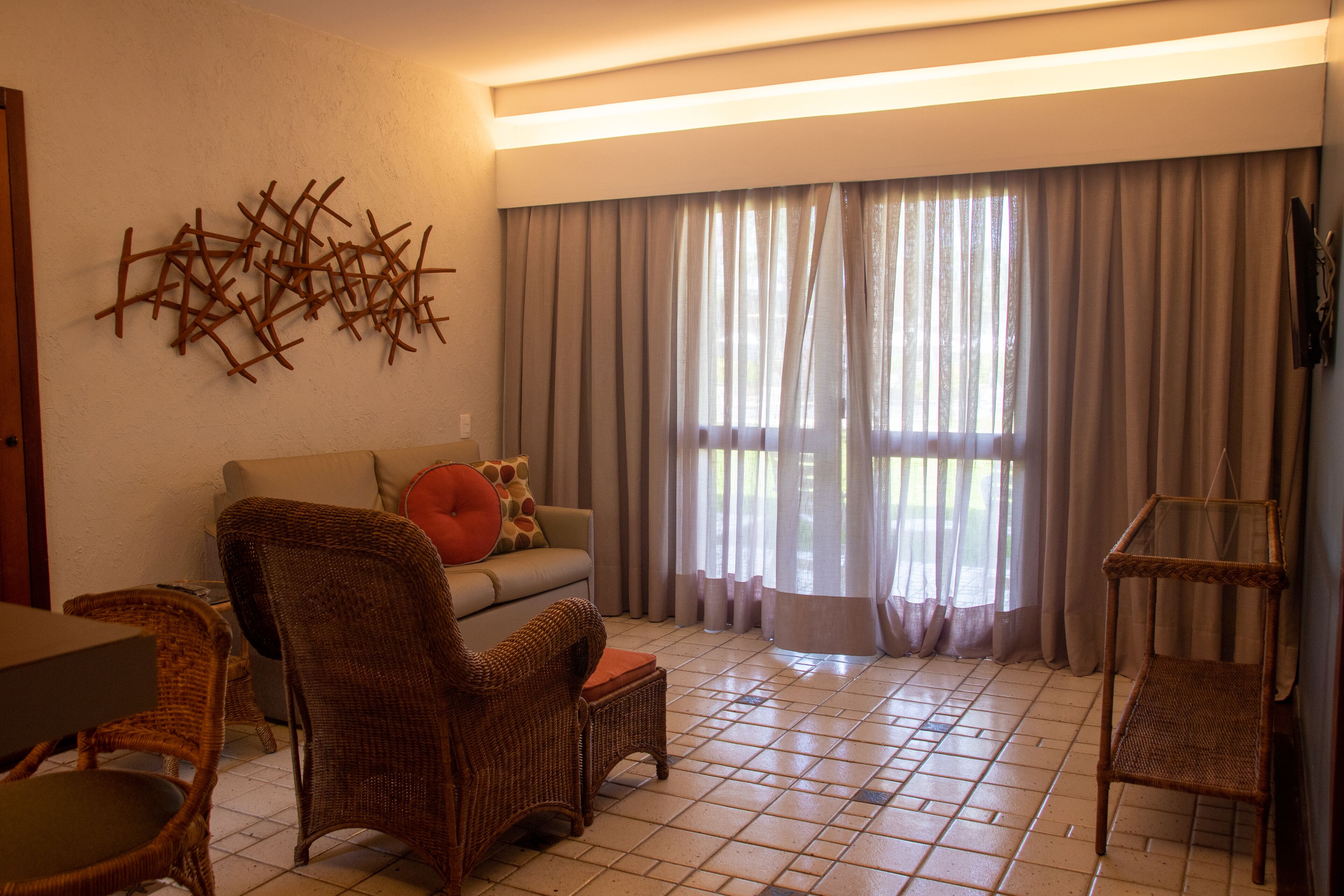 Una sala de estar con un sofá, dos sillones de mimbre, una mesa de centro de vidrio y un televisor instalado en la pared.