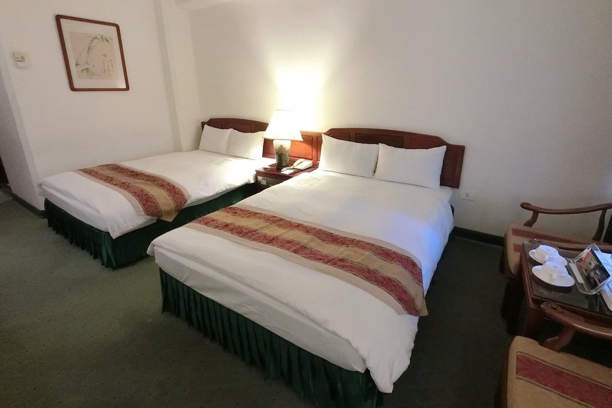 A hotel room with two beds, a nightstand with a lamp, a framed picture on the wall, and a small table with cups and a book.