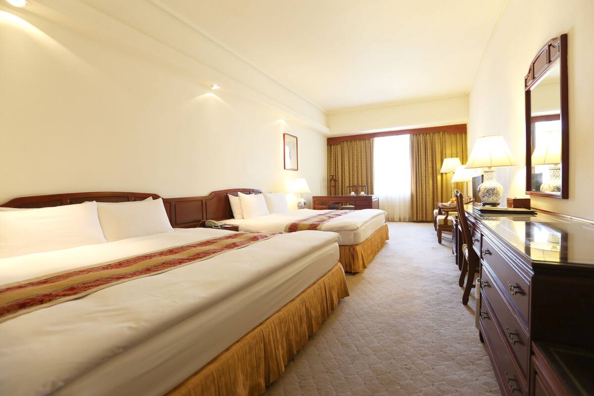 A hotel room with two beds, a dresser, a desk, and a large window with curtains.