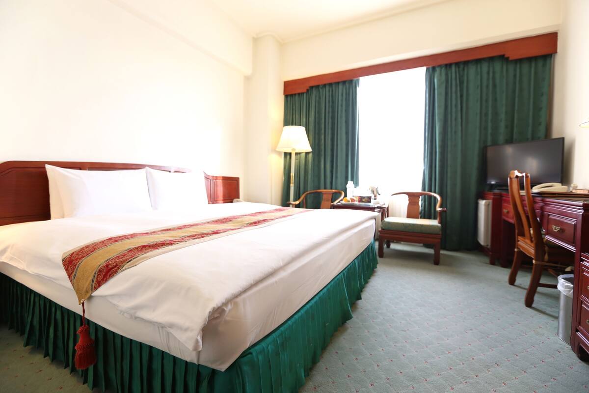 A hotel room with a large bed, a desk, a chair, a television, and a window with curtains.