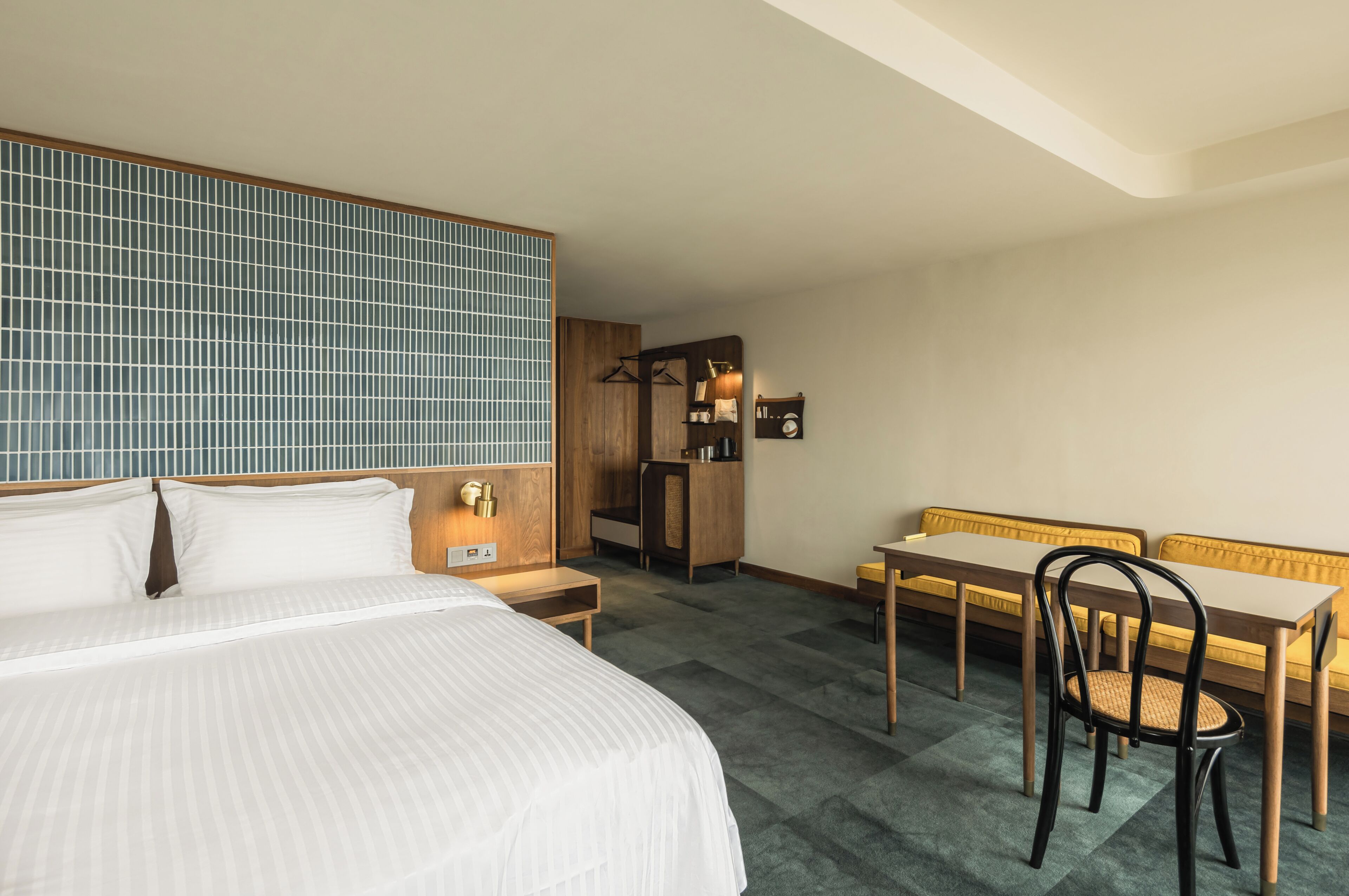 A hotel room with a bed, a wooden desk, a chair, a bench, and a wardrobe.