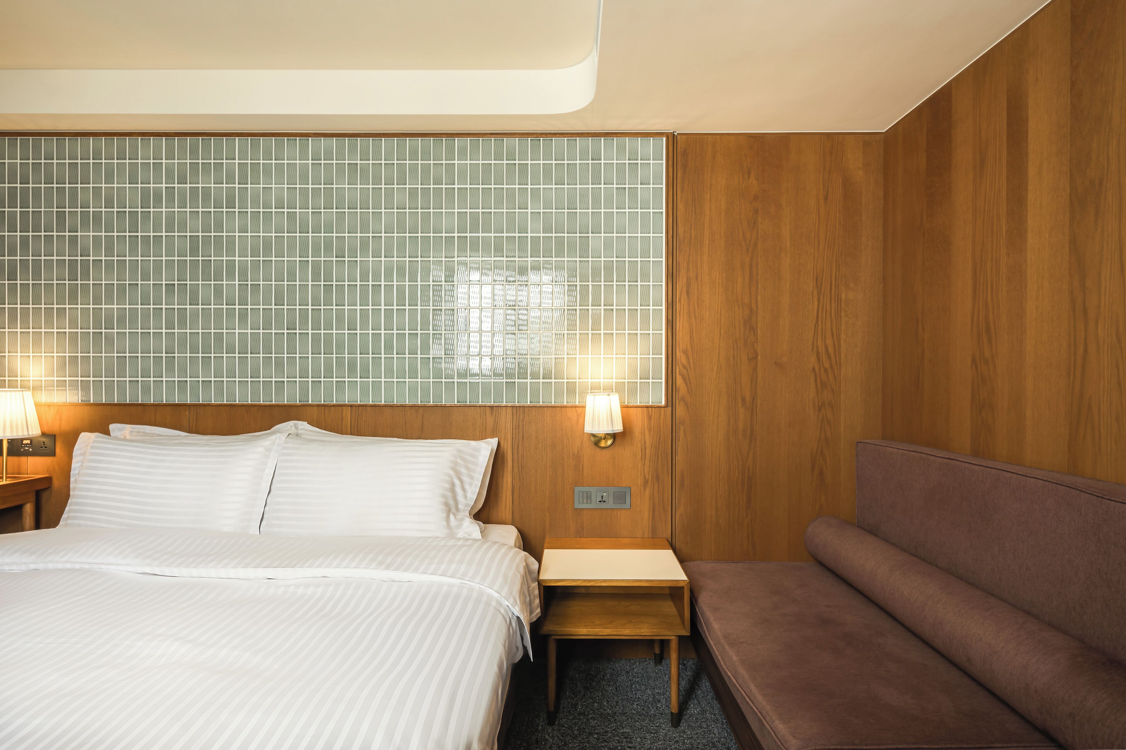 A hotel room with a large bed, a wooden headboard, a small bedside table with a lamp, and a bench.