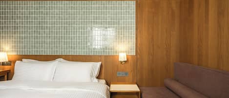 A hotel room with a large bed, a wooden headboard, a small bedside table with a lamp, and a bench.