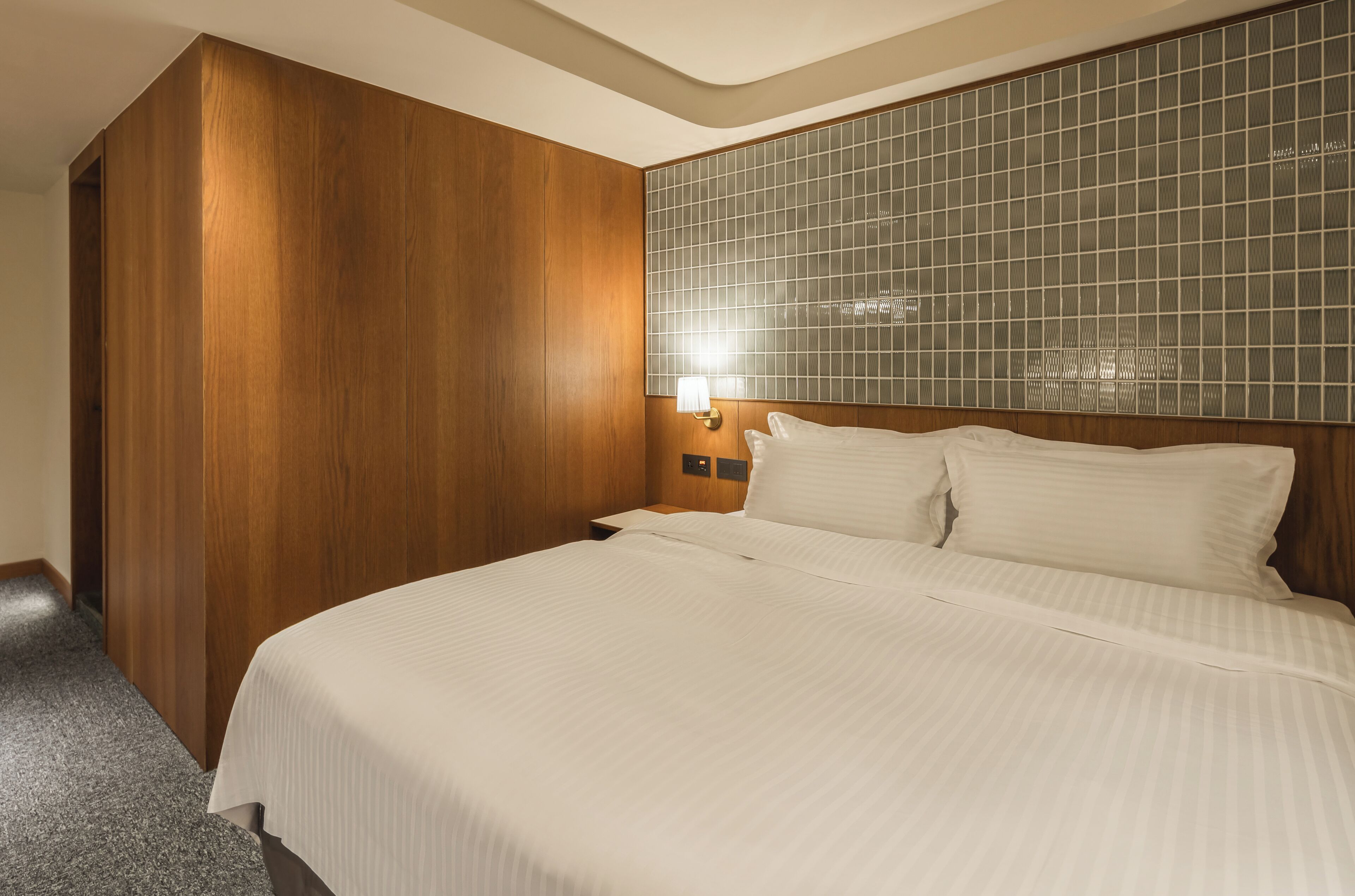 A hotel room with a large bed, wooden panel walls, a tiled headboard, and a nightstand with a lamp.