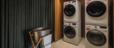 Une buanderie équipée de deux machines à laver à chargement frontal superposées, d’un seau en métal posé sur un support en bois et d’un rideau vert foncé.