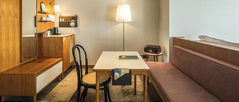 A compact hotel room with a wooden desk, a bench, a chair, a bedside table, a lamp, and a small radio.