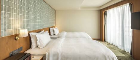 A hotel room with two beds, a bedside table with a radio, a wall-mounted TV, and a large window with curtains.