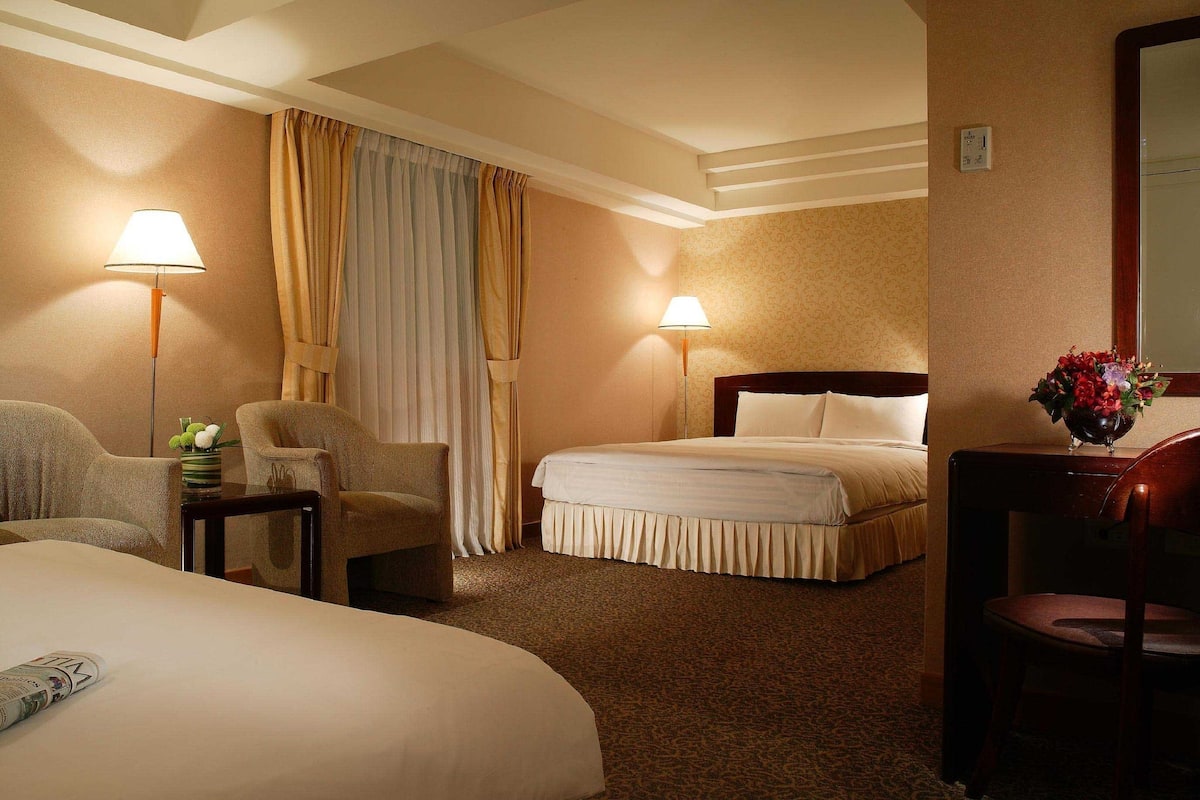 Una habitación de hotel con una cama grande, dos sillones, una mesita con una lámpara y una mesita de noche.