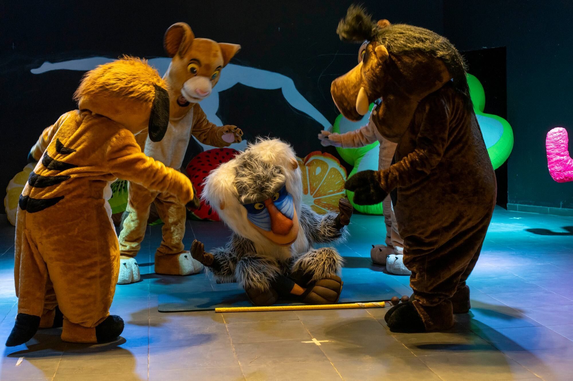 A group of people in animal costumes on a stage with a blue floor and a large green and pink inflatable structure in the background.