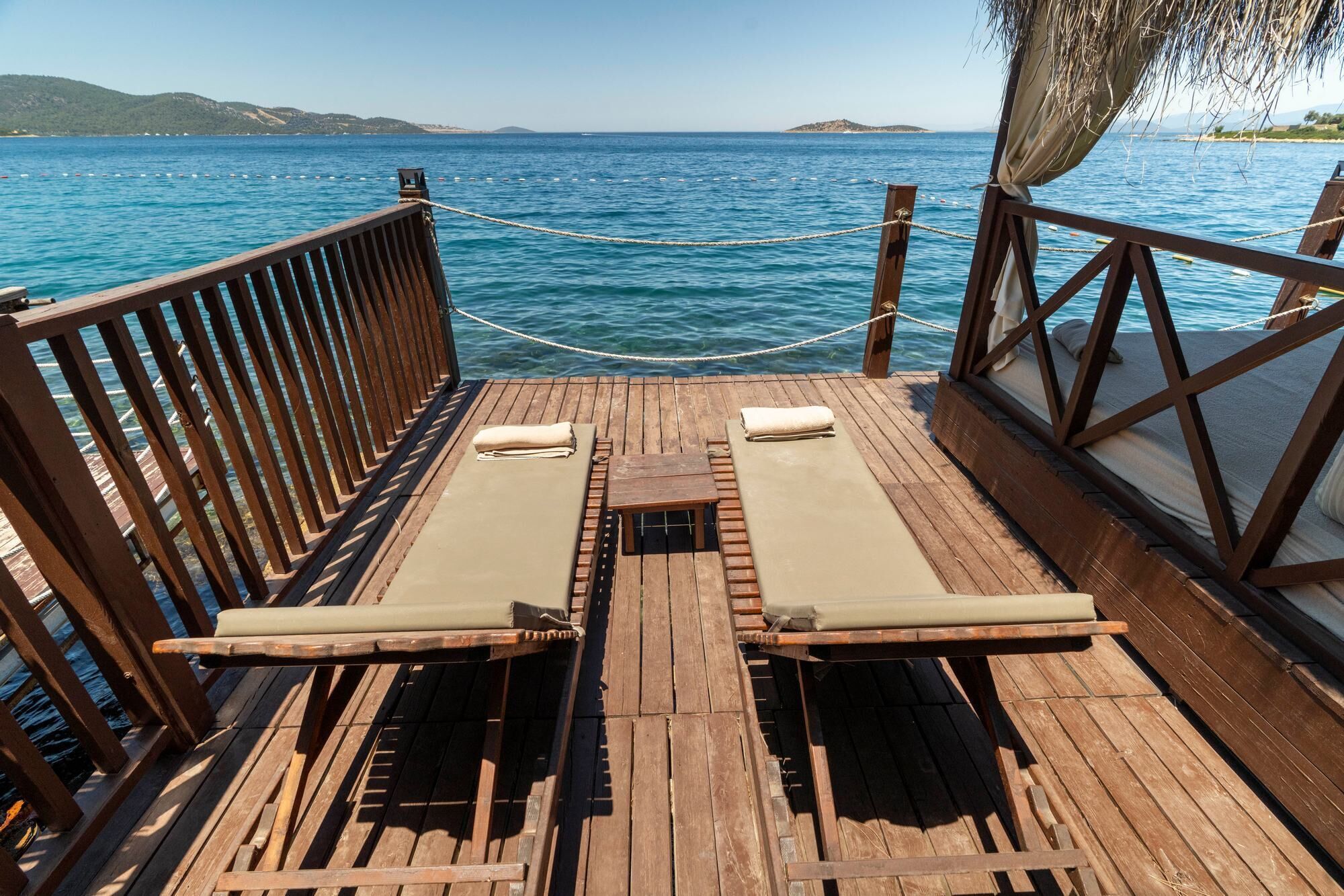 A wooden deck with two lounge chairs and a small table, overlooking a calm sea with buoys and distant hills.