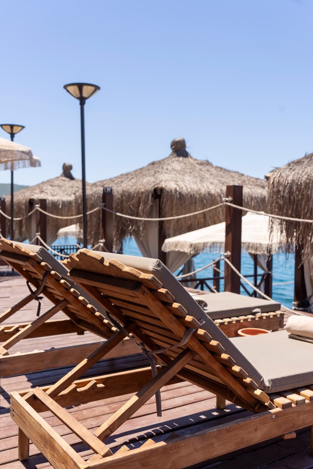 Wooden lounge chairs with cushions on a wooden deck, thatched umbrellas, and a clear view of the sea.