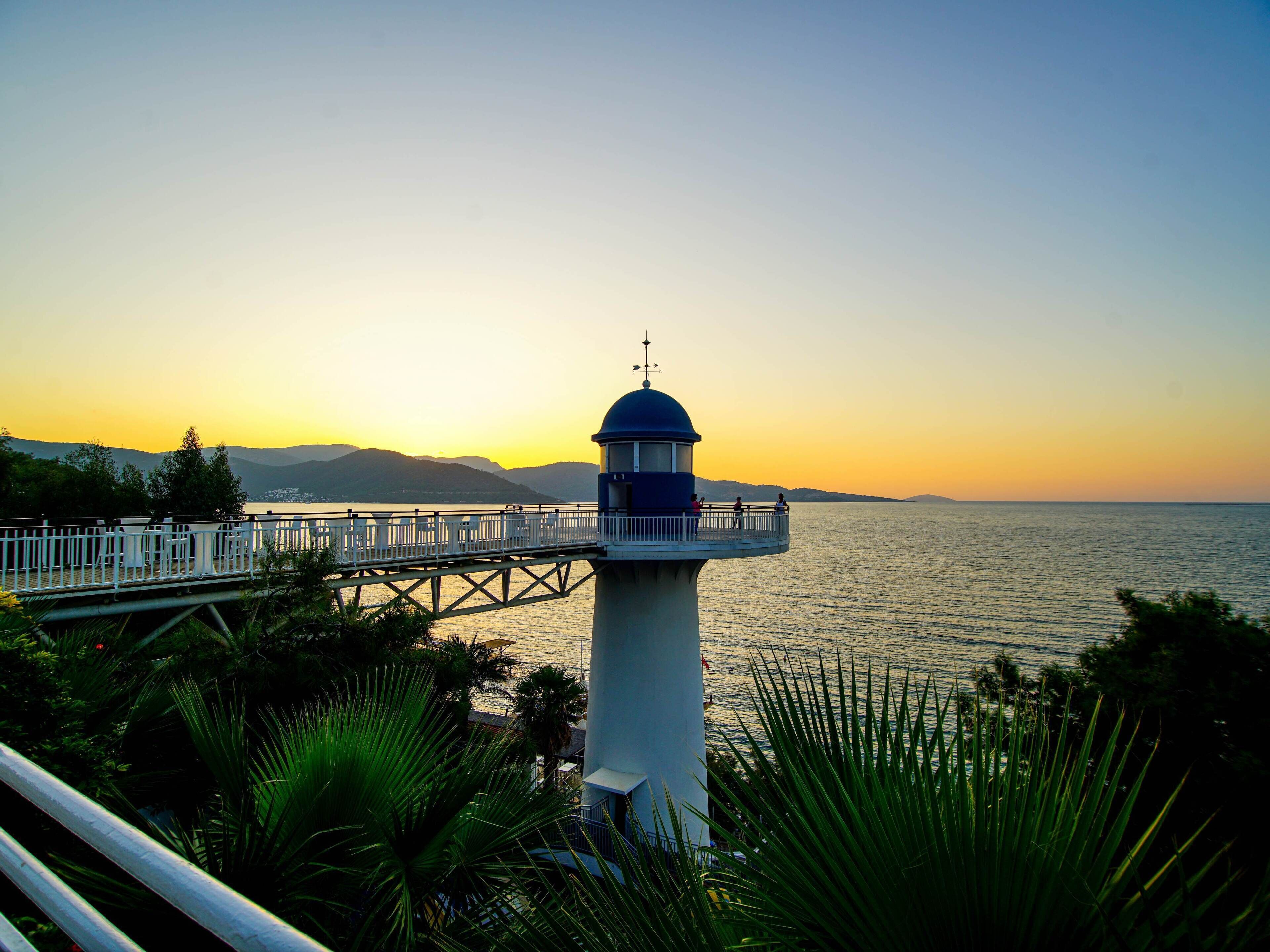 A lighthouse with a white and blue structure, a bridge leading to it, and a view of the sea and mountains in the background.