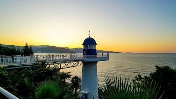 A lighthouse with a white and blue structure, a bridge leading to it, and a view of the sea and mountains in the background.
