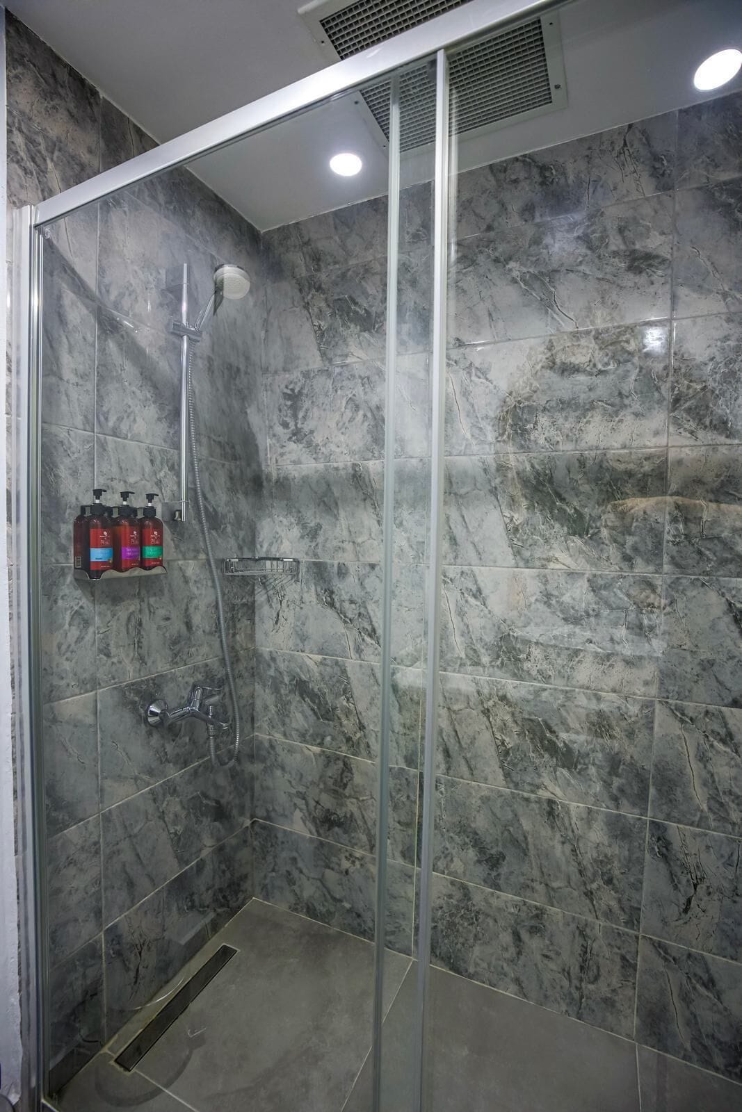Une douche fermée par des parois de verre, avec un mur en marbre, une pomme de douche à main et un distributeur de savon à main.