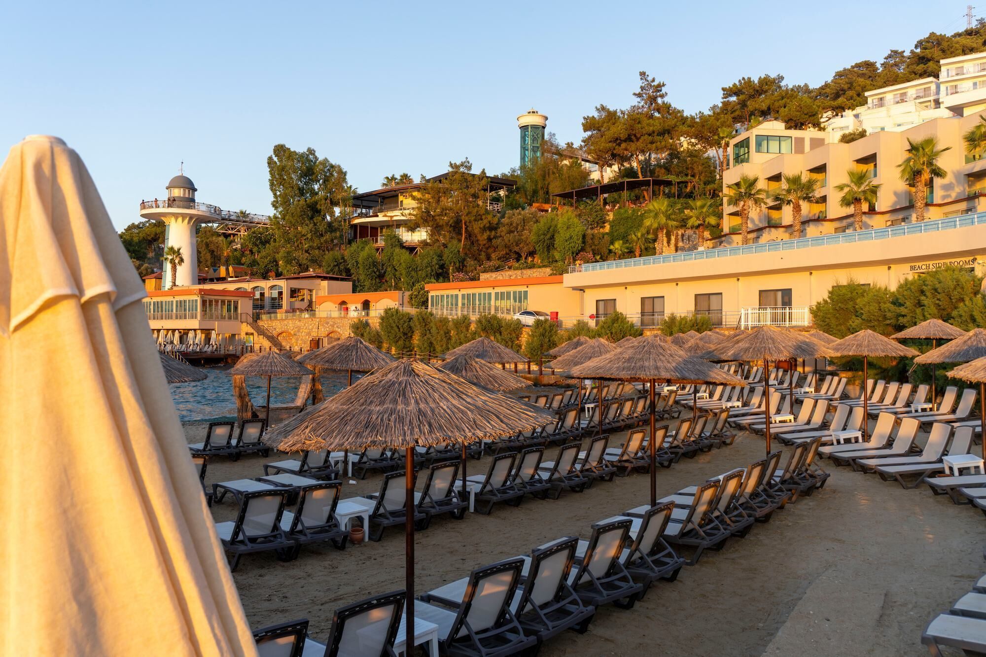Un hotel frente a la playa con hileras de tumbonas y sombrillas de paja, un faro y una torre al fondo.