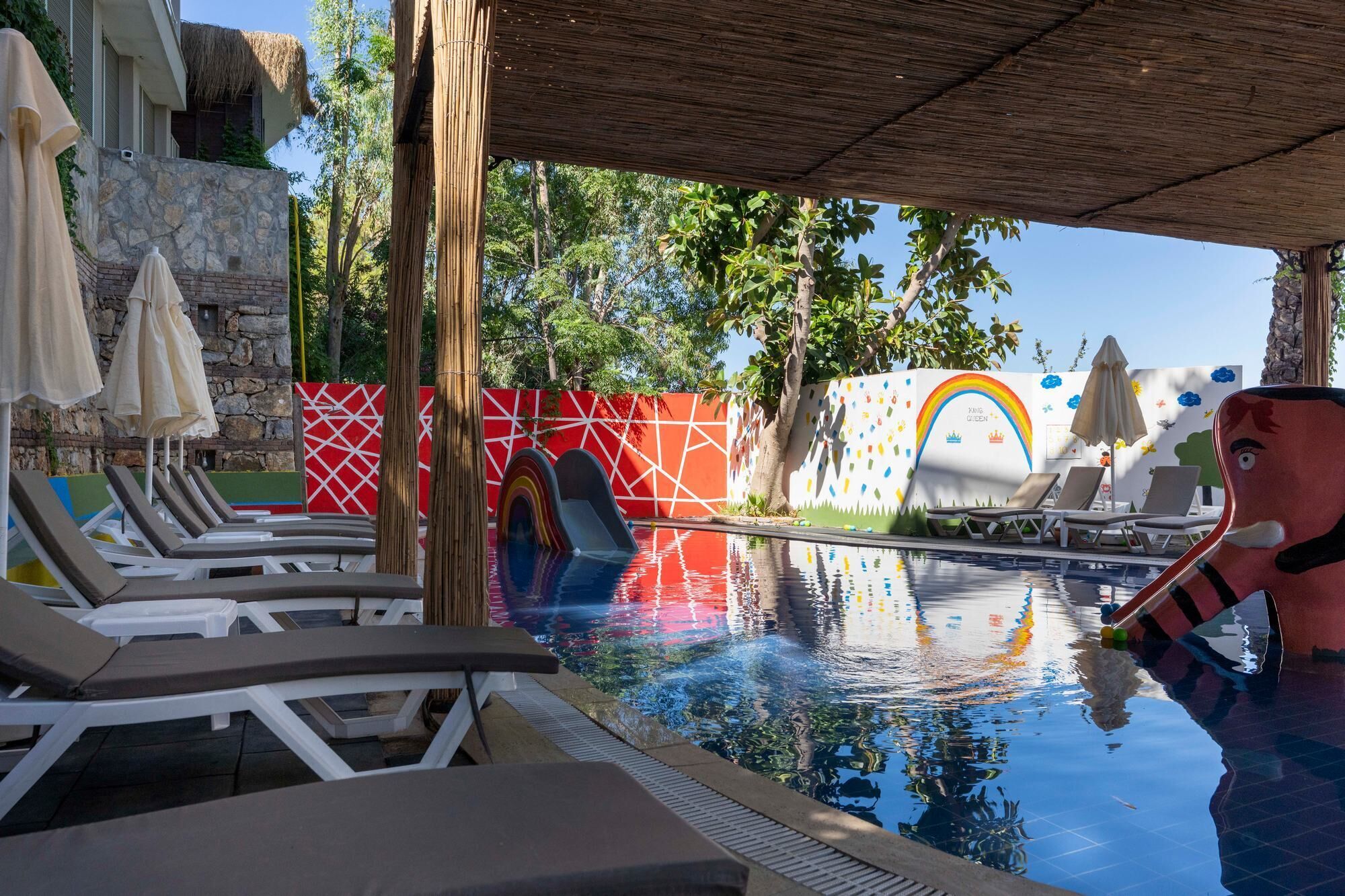 A swimming pool with lounge chairs and umbrellas, surrounded by a stone wall and colorful murals.