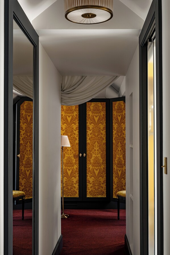 A hallway with a red carpet, a floor lamp, and a mirror reflecting a room with patterned wallpaper and a chair.