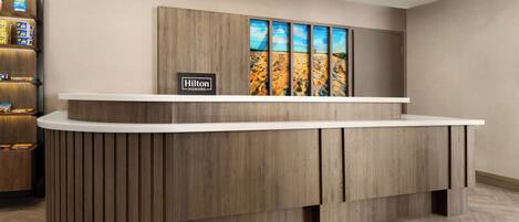 Ein Empfangsschalter mit Holzoptik, ein Schild mit der Aufschrift "Hilton Honors" und ein Kunstwerk zum Thema Wüste darüber.