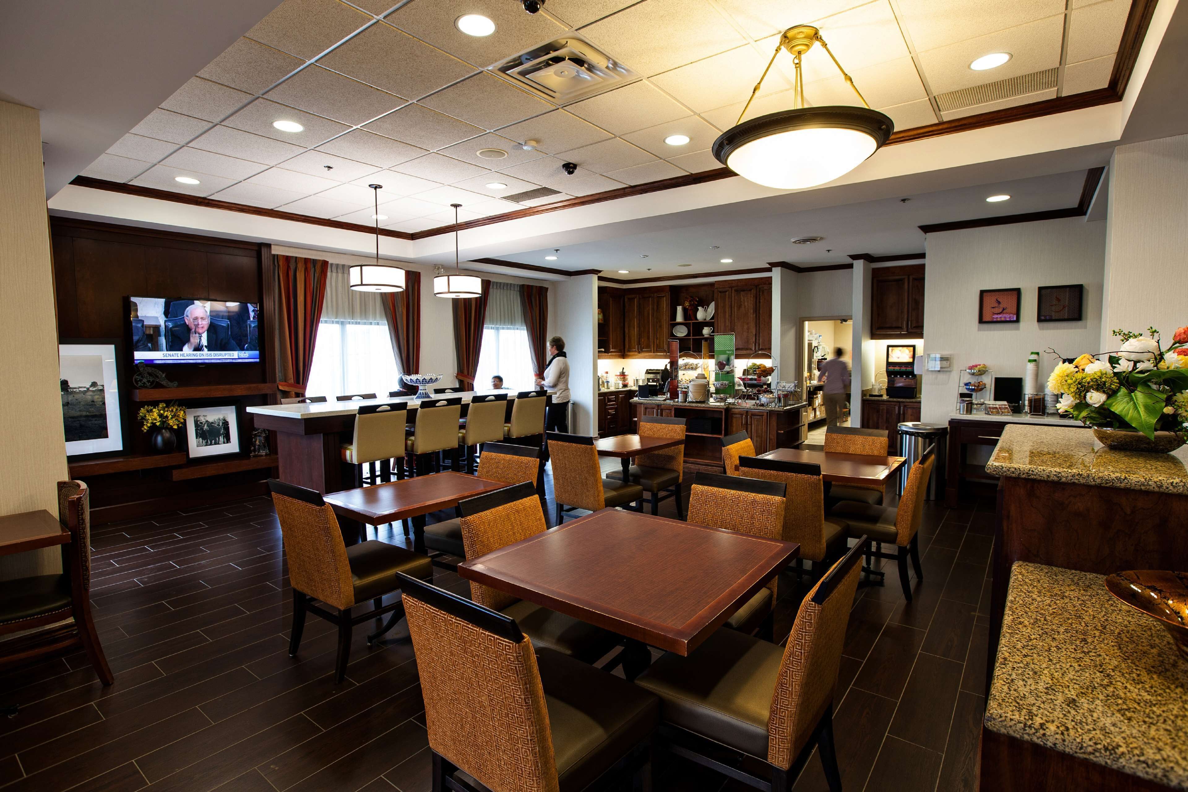 A hotel dining area with multiple tables and chairs, a television screen, and a kitchen in the background.