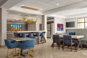 A modern dining area with a long table and blue chairs, a bar area with stools, and a large TV on the wall.