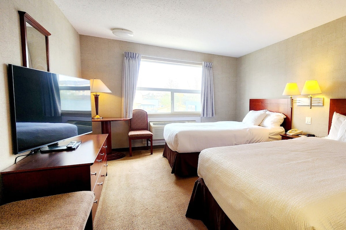 A hotel room with two beds, a TV, a desk, a chair, and two lamps.