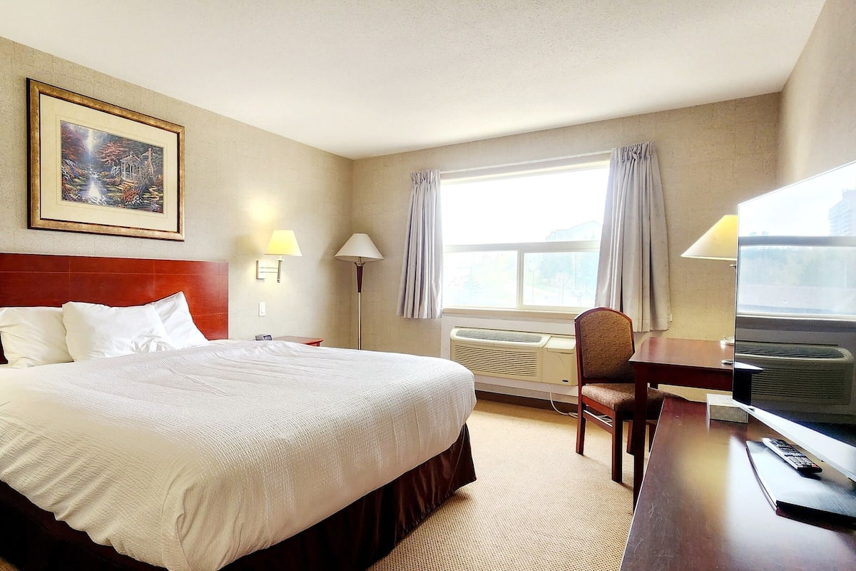 A hotel room with a large bed, two bedside lamps, a desk with a TV, a chair, and a window with curtains.