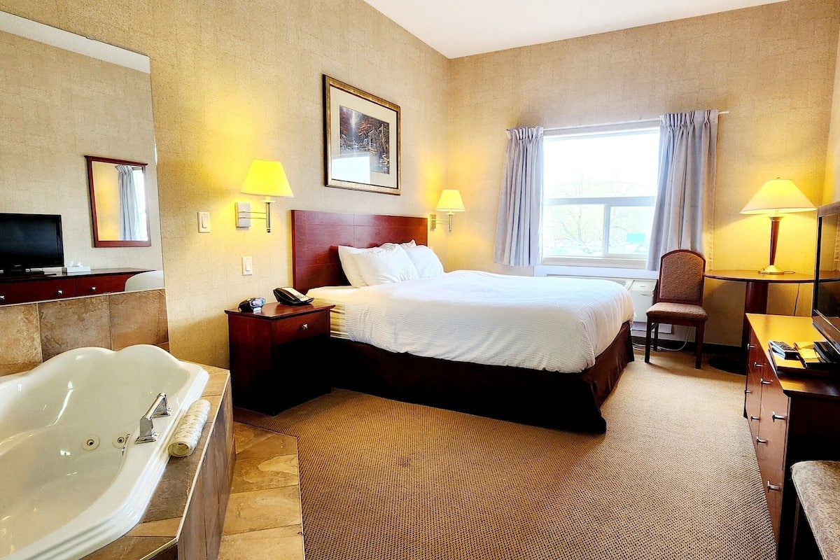 A hotel room with a large bed, a sitting area with a chair, a desk, a television, and a bathtub.