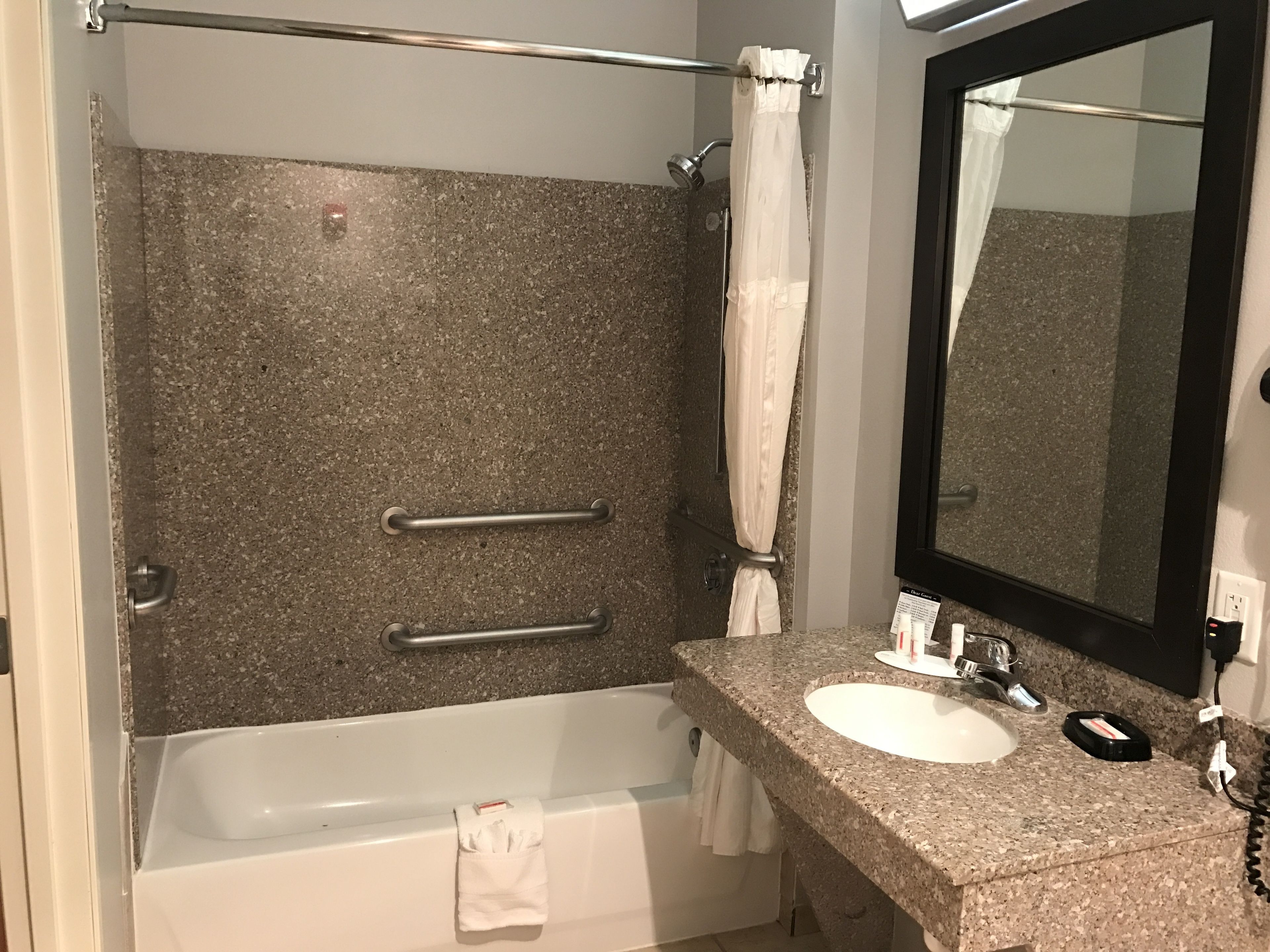 A bathroom with a granite countertop, a white sink, a mirror, a shower with a curtain, and grab bars.