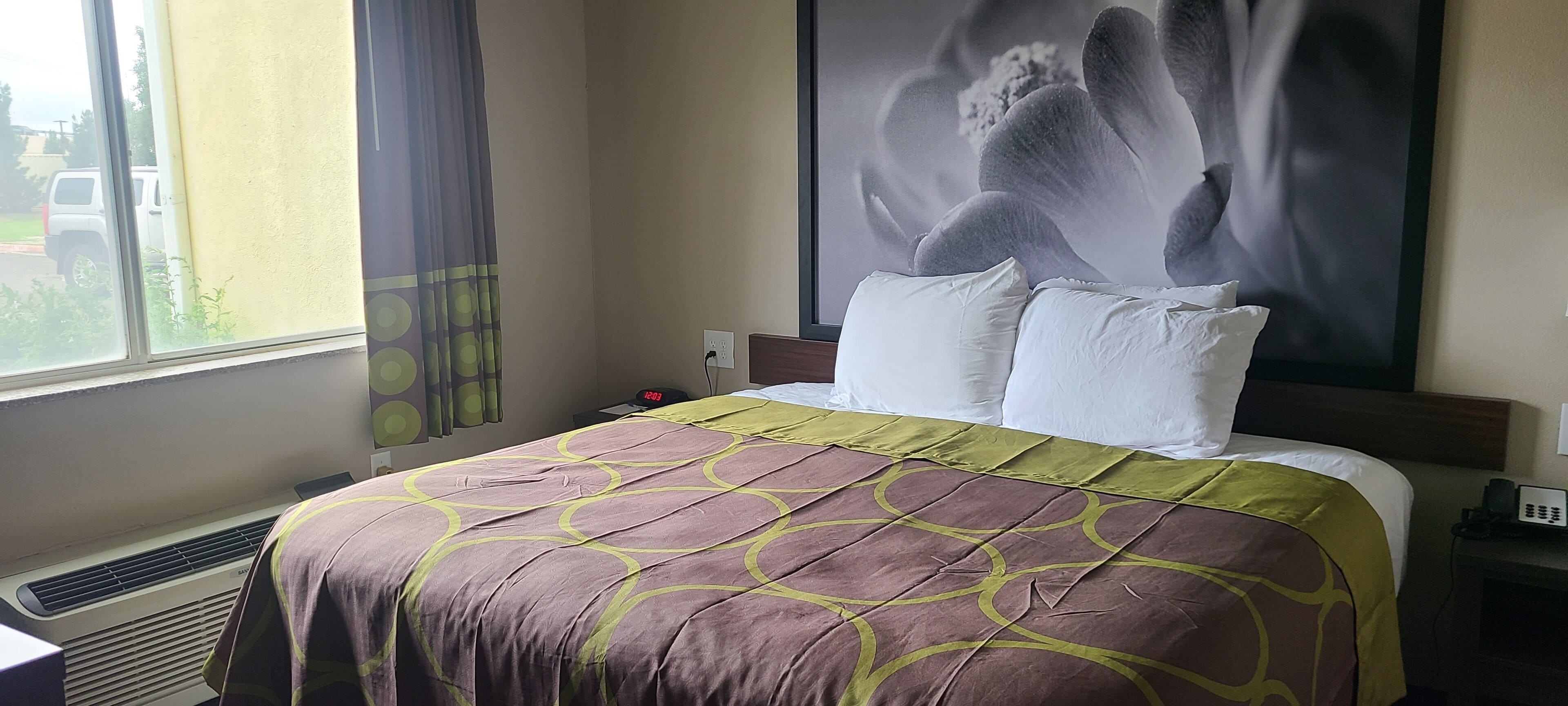 Habitación de hotel con cama, mesita de noche, reloj de pared y ventana con cortinas.