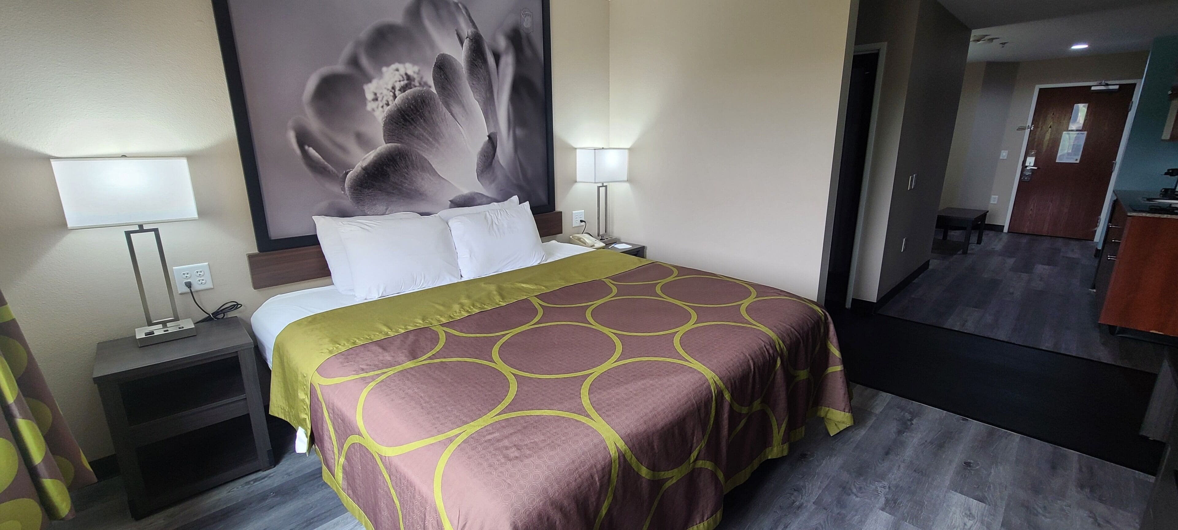 Une chambre d’hôtel avec un lit, deux lampes de chevet, une table de chevet et une grande œuvre d’art florale au mur, donnant sur le couloir.