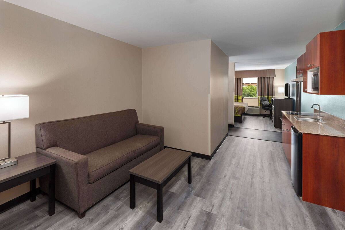 Hotelkamer met een sofa, een klein tafeltje, een kitchenette met een koelkast en een raam met gordijnen.