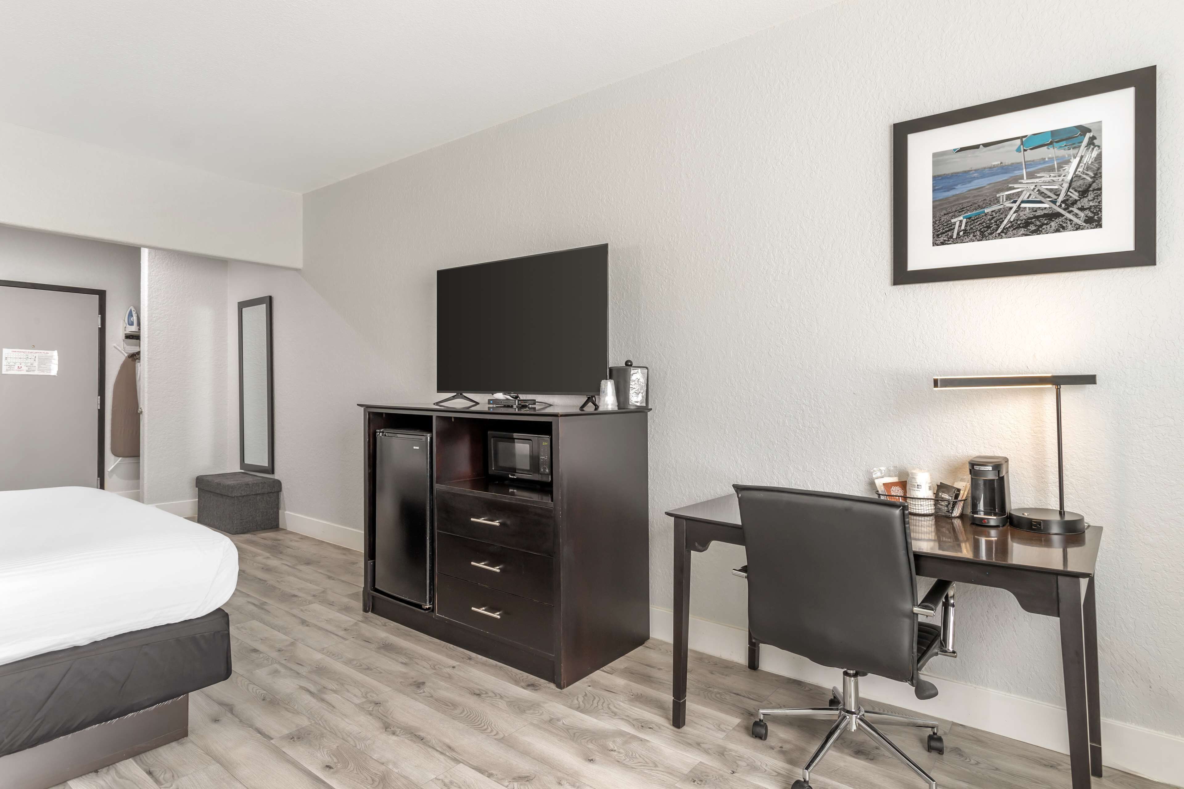 Habitación de hotel con cama, escritorio, cómoda oscura, televisor de pantalla plana, un cuadro enmarcado en la pared y un pequeño cesto de basura.