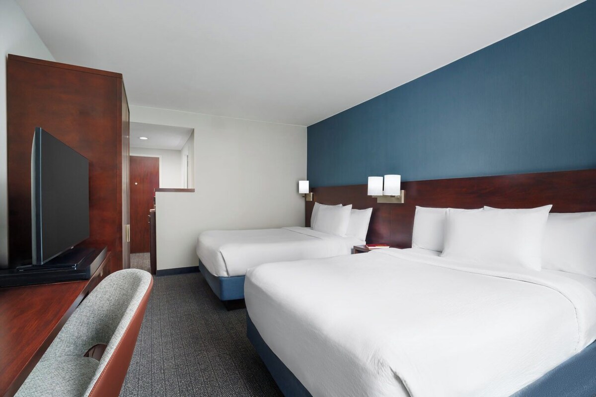 Ein Hotelzimmer mit zwei Betten, einem Flachbildfernseher, einem Schreibtisch und einem Sessel.
