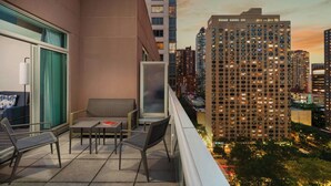 Un balcón con sofá, sillas y una mesa, con vistas a un paisaje urbano con rascacielos al atardecer.