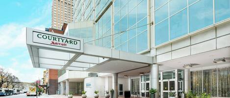 L’entrée d’un hôtel Courtyard Marriott, avec une allée couverte, de grands pots de plantes et un ciel dégagé.